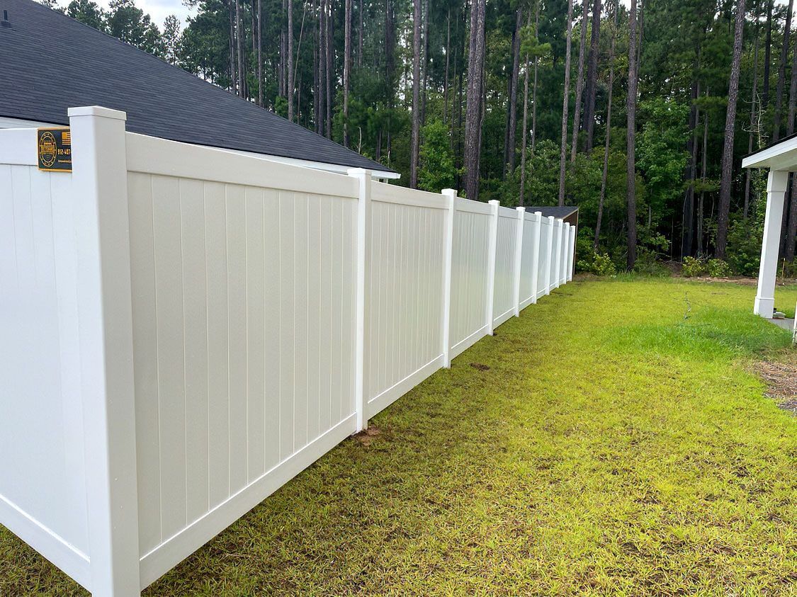 White vinyl privacy fence along a grassy yard beside a house, with trees in the background