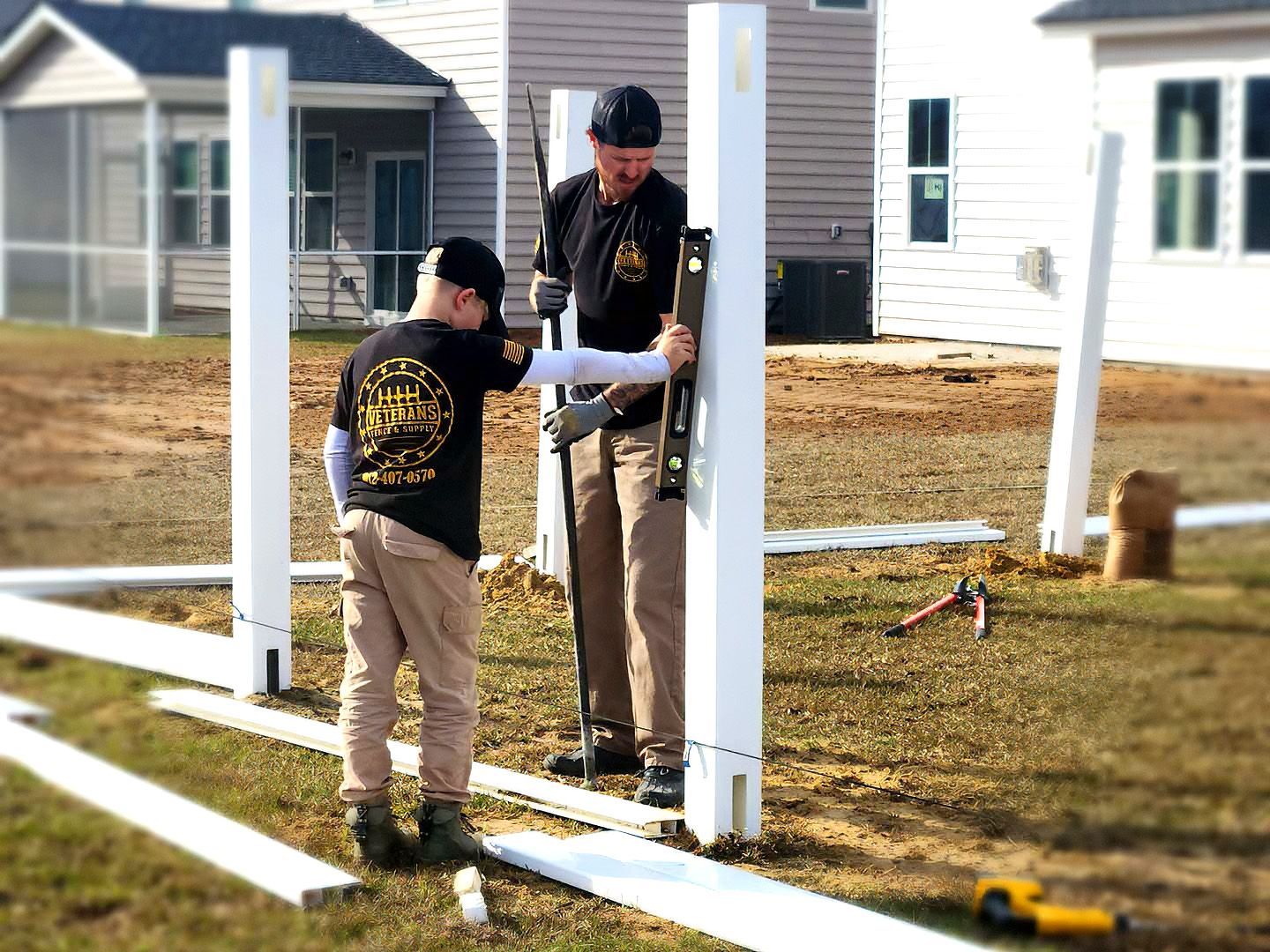 Two workers assembling white fence posts in a suburban yard under construction.
