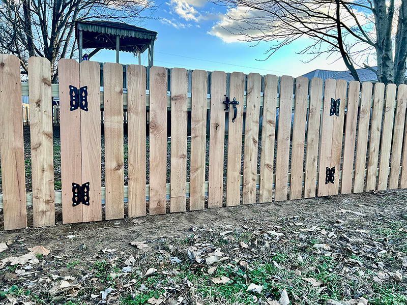 Wooden fence with black butterfly decorations in a yard, with trees and a blue sky behind it.