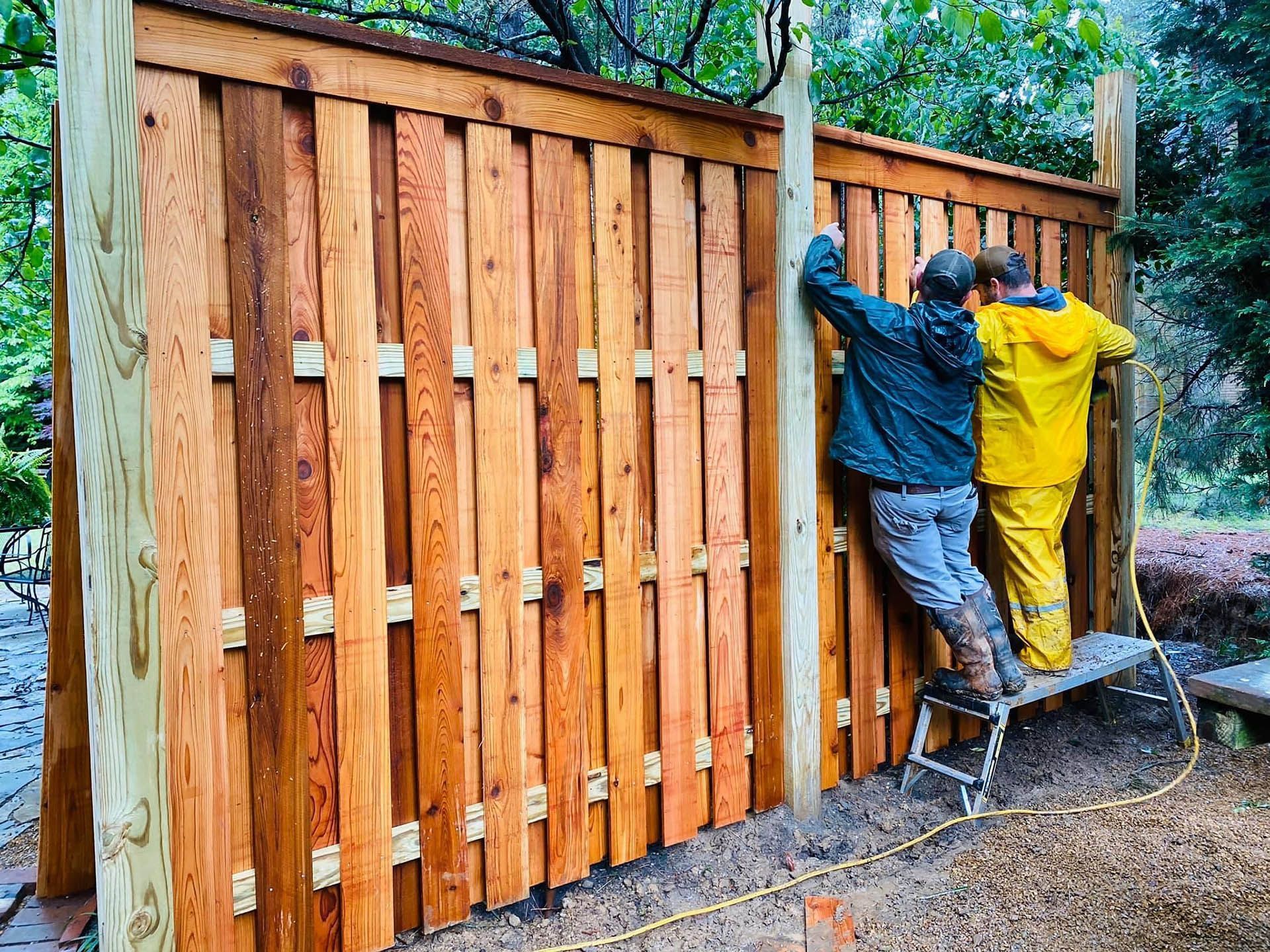 Two workers install a wooden fence panel outdoors, one in blue and one in yellow.