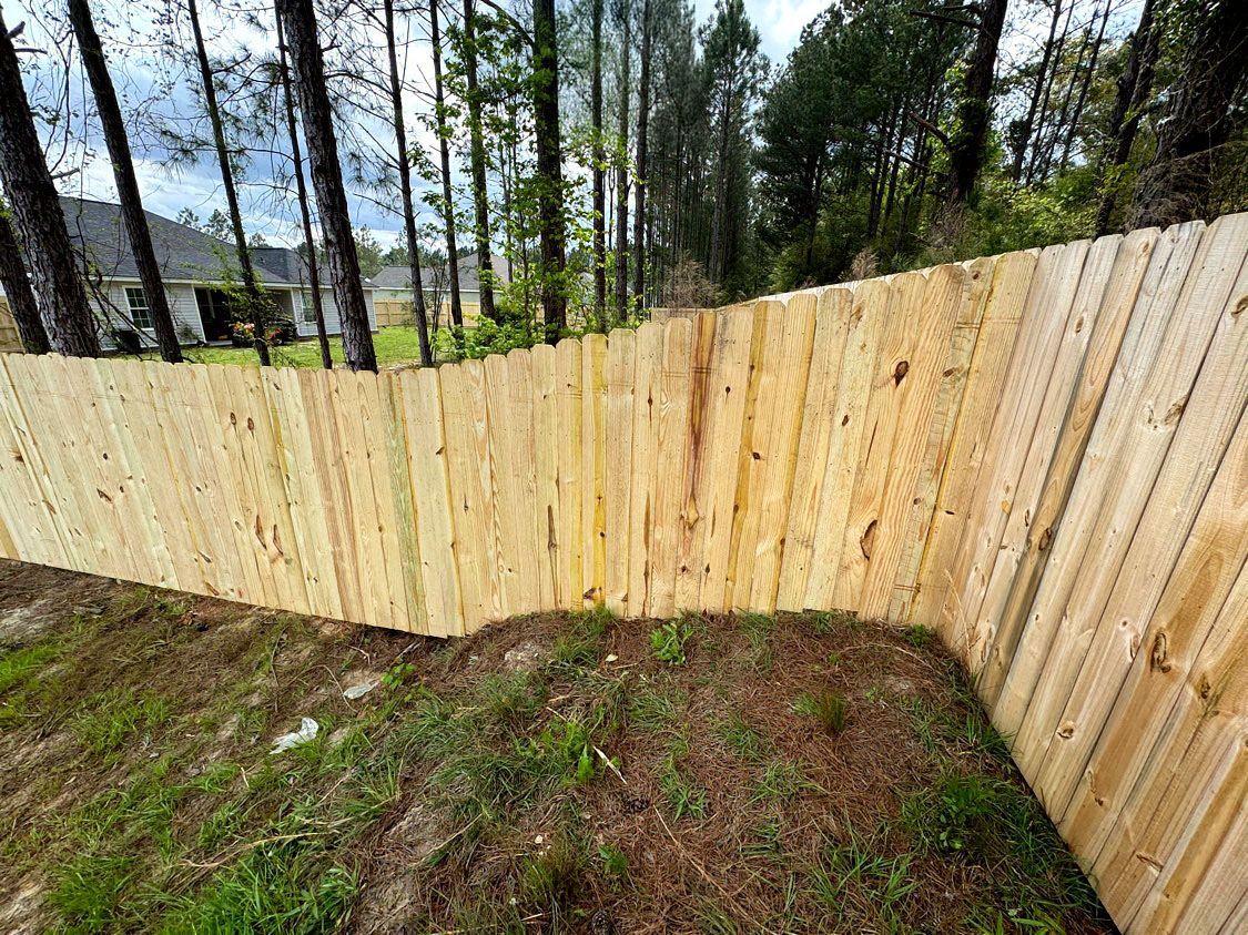 New wooden fence enclosing a dirt yard with trees and a house in the background
