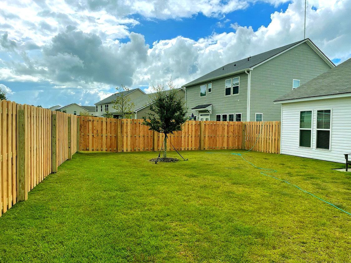 Backyard with green lawn, wooden fence, young tree, and cloudy sky beside a house