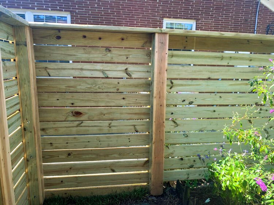 Wooden slat privacy fence in a sunny yard with a brick wall behind it and shrubs at the right edge