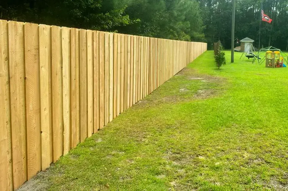 Long wooden fence along a grassy yard beside trees and a small playground in the distance