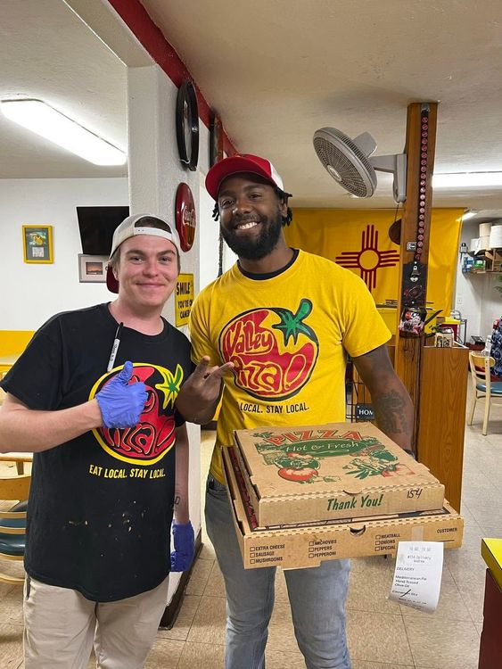 Two smiling people in a pizza shop holding a boxed pizza and wearing colorful T-shirts.