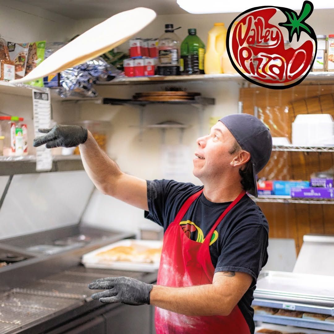 Chef tossing dough in a busy pizzeria kitchen, wearing a red apron and black gloves.
