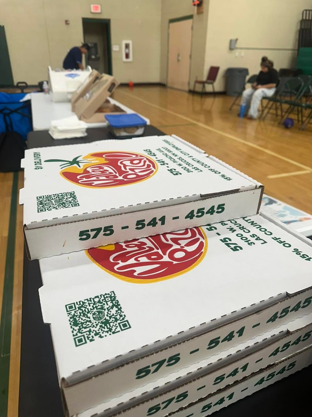 Stacked pizza boxes on a gym floor with people seated in the background