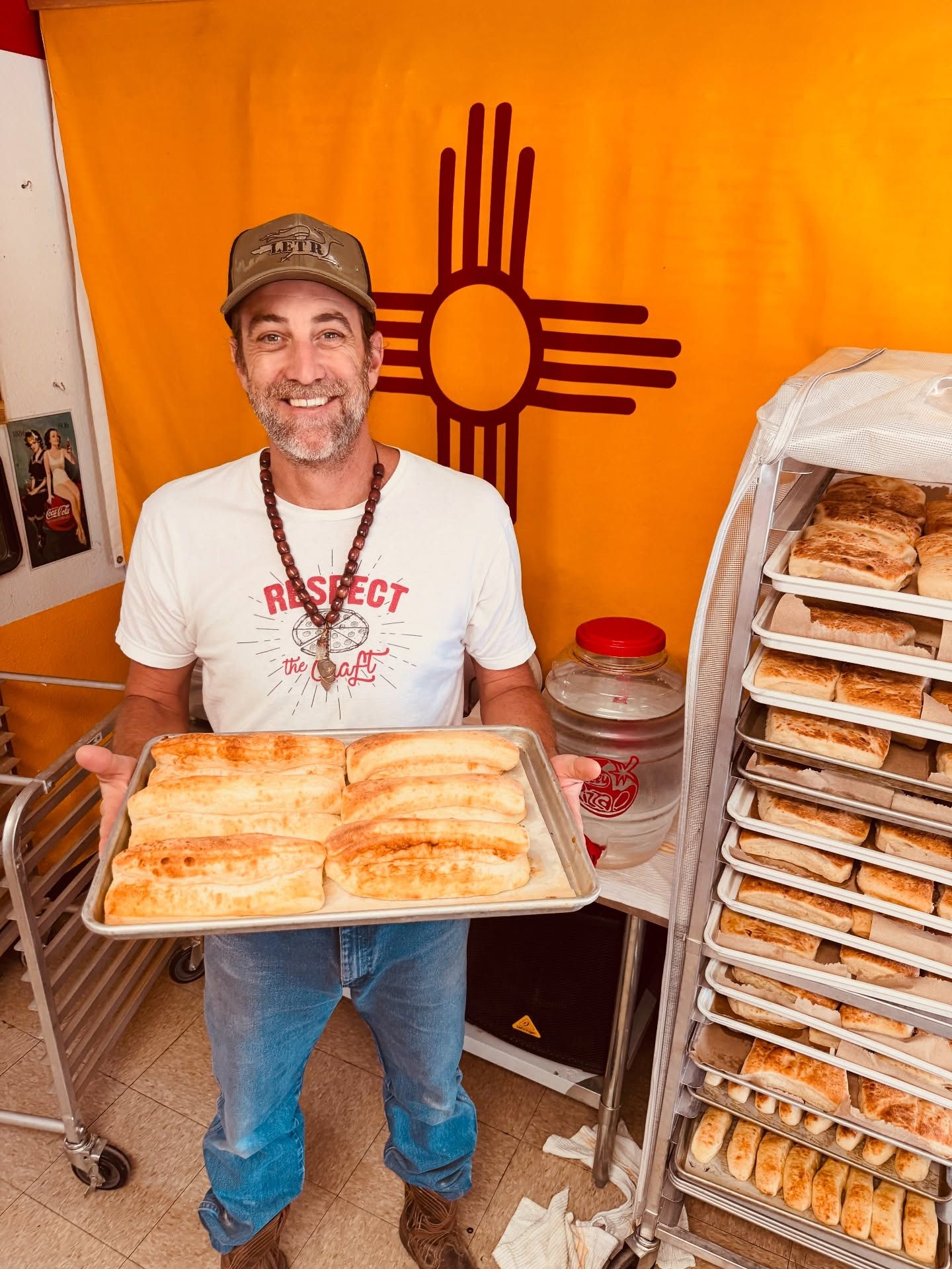Man holding a tray of baked pastries in front of an orange New Mexico flag and a rack of bread loaves.