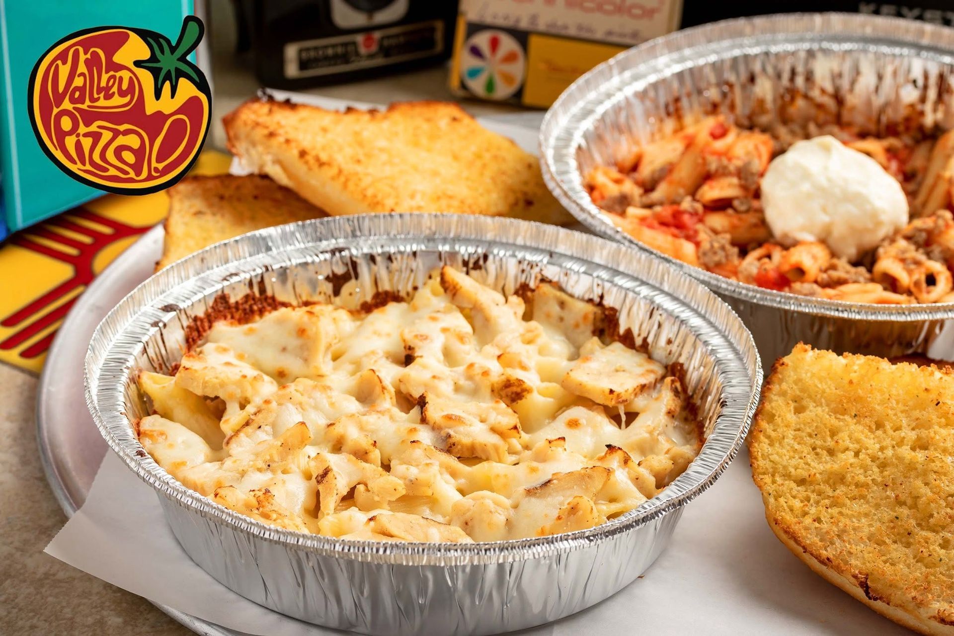 Two foil pans of cheesy baked pasta with bread slices on a table, with a logo in the corner
