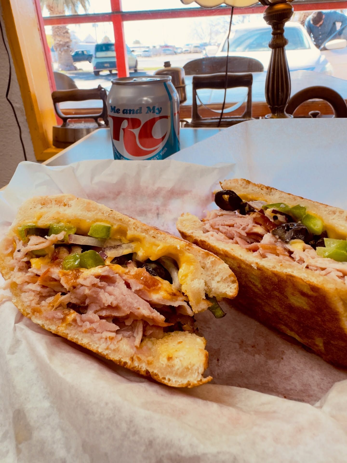 Two halves of a deli sandwich on paper, with a Dr Pepper can and a window view in the background