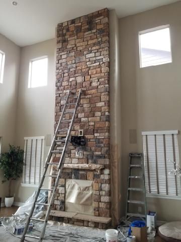 A tall stone fireplace under construction, with ladders and tools in a room featuring multiple windows and beige walls.