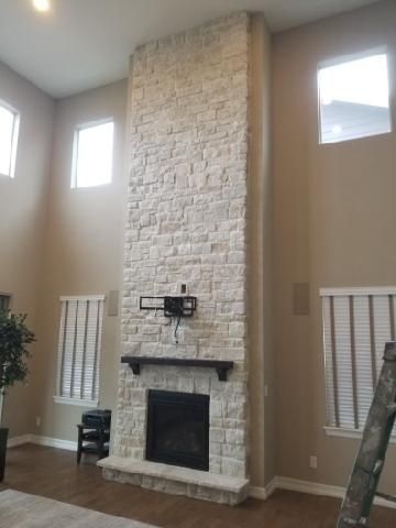 A tall, light-colored stone fireplace chimney extends to a vaulted ceiling in a beige living room with two windows.