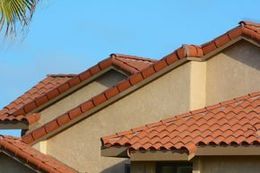 A close-up of a house roof featuring red clay tiles and beige stucco walls against a clear blue sky.