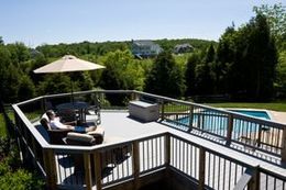 A person relaxes on a chair on a multi-level deck overlooking a swimming pool and wooded landscape.