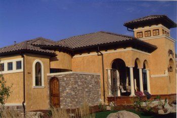 A single-story, tan stucco Mediterranean-style house with a tile roof, a small stone-faced wall, and a decorative tower.