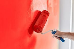 A hand uses a paint roller to apply vibrant red paint to a wall.