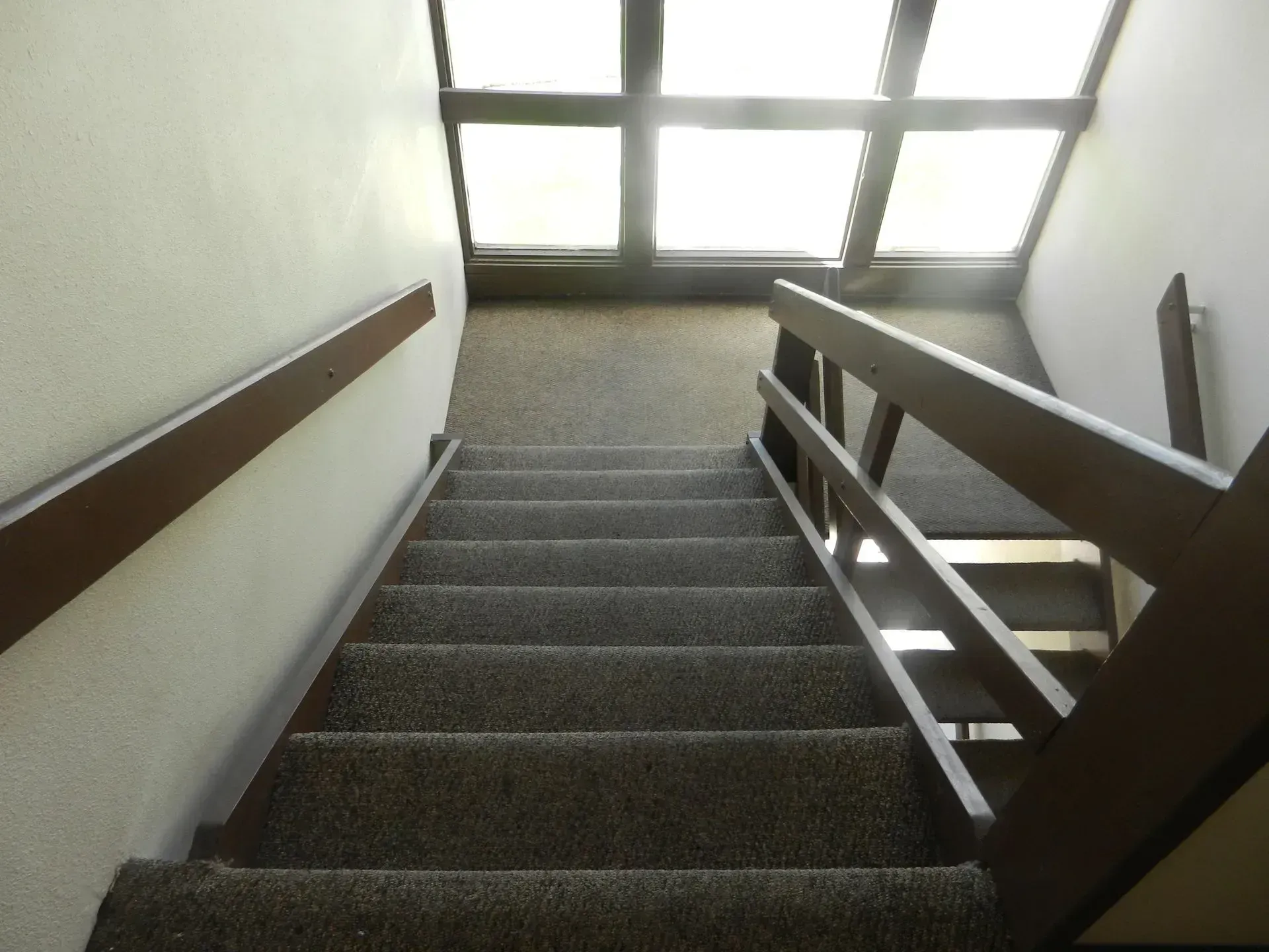 Looking down a carpeted staircase with wooden handrails toward a landing with a large window.