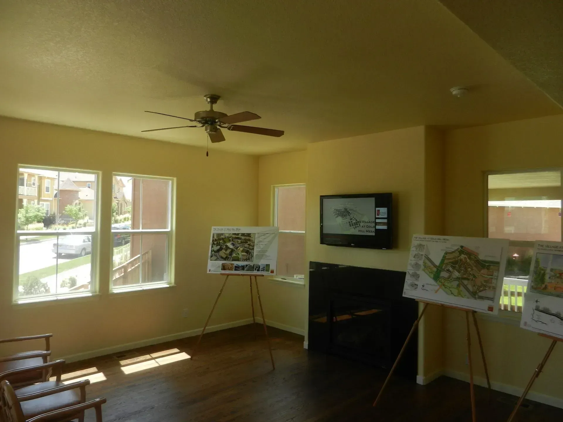 A yellow-walled room with two presentation boards on easels, a ceiling fan, and a flat-screen TV mounted on the wall.