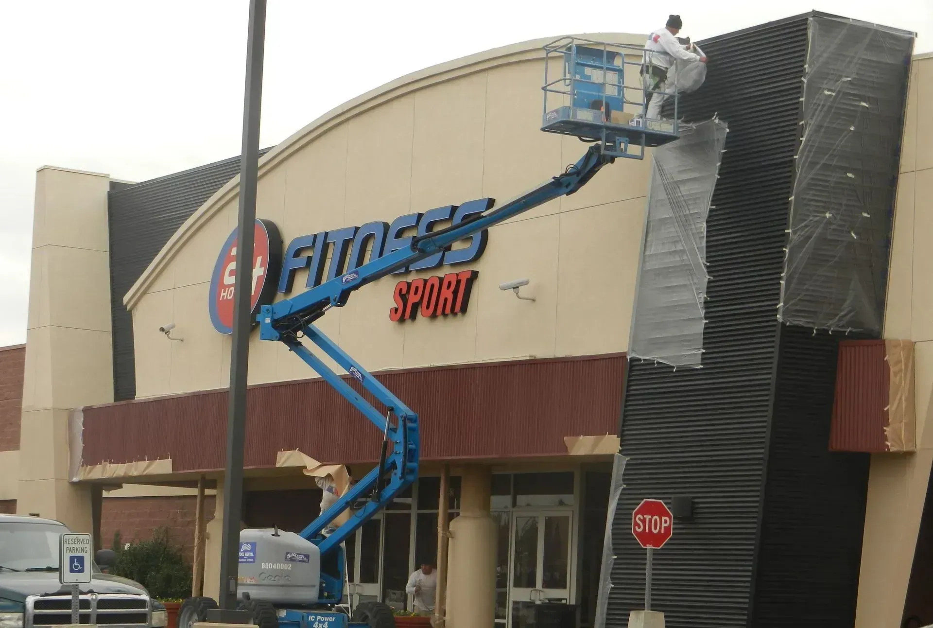 A worker in a boom lift repairs the exterior wall and logo of a 24 Hour Fitness Sport gym building.
