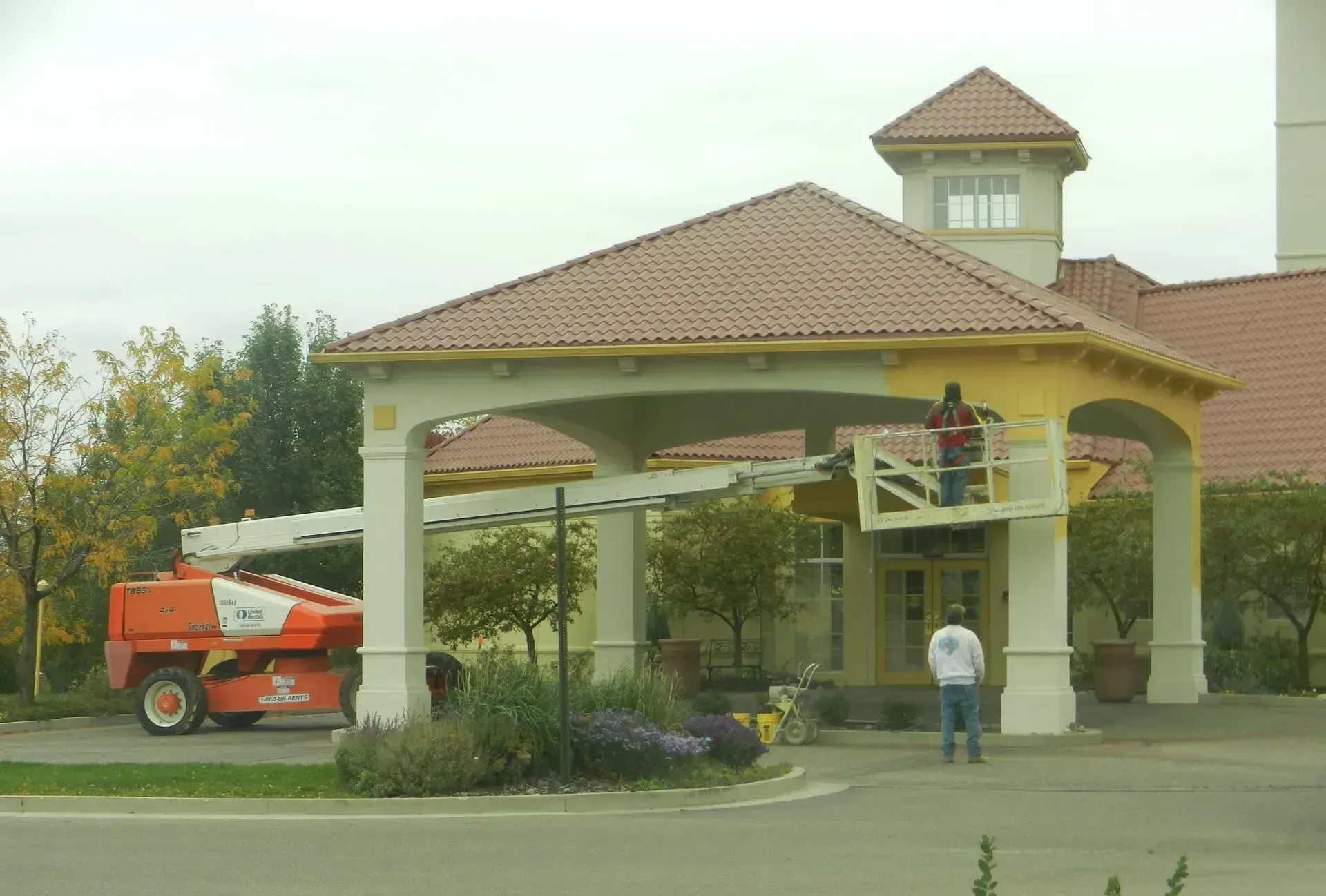 A worker uses an aerial lift to paint the trim of a building's entrance canopy while another person stands nearby.