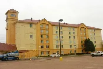 A four-story, yellow hotel building with a clock tower and red roof sits near a parking lot under a cloudy sky.