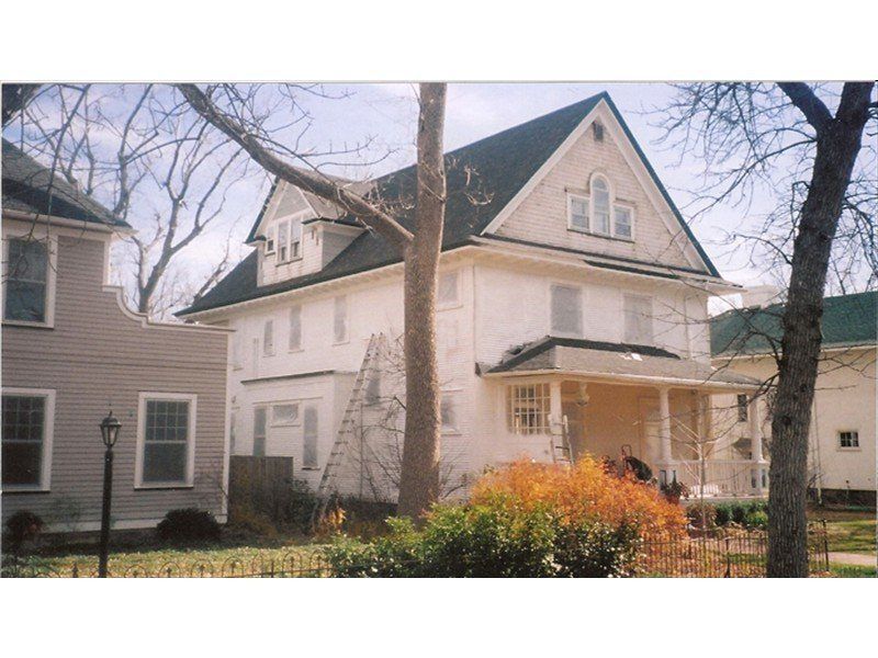 A white two-story house with a green roof, gables, and a front porch, flanked by trees and neighboring houses.