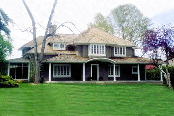 A two-story dark-colored house with a large front porch, surrounded by green lawns and trees under a cloudy sky.