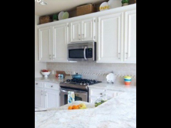 A bright kitchen with white cabinets, stainless steel appliances, a marble countertop, and decorative items above shelves.