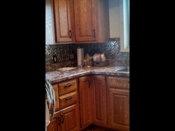 A corner kitchen view with medium-toned oak cabinets, patterned laminate countertops, and a dark, embossed backsplash.