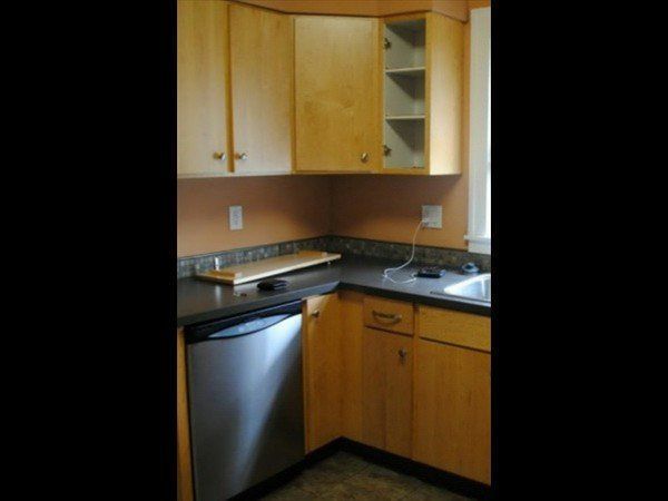 A kitchen corner featuring light wood cabinets, dark countertops, a stainless steel dishwasher, and an open corner shelf.