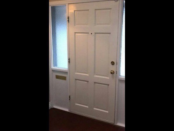 A white, six-panel front door with brass hardware and a side glass panel, set against a white wall and dark floor.
