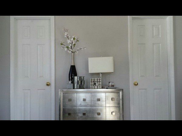 A silver dresser sits between two closed white doors, topped with a lamp, a vase of flowers, and small decor items.