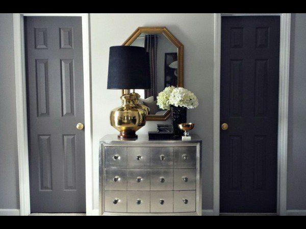 A silver chest sits between two dark grey doors, topped with a brass lamp, a mirror, and a vase of white flowers.
