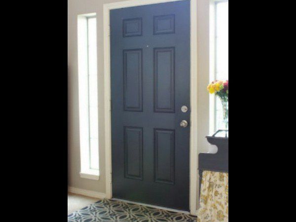 A dark-colored, six-panel front door flanked by vertical side windows, with a patterned rug and a vase of flowers nearby.
