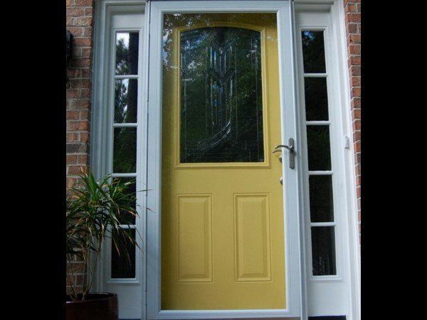 A yellow front door with a decorative glass window panel, framed by white trim and vertical side windows on a brick house.