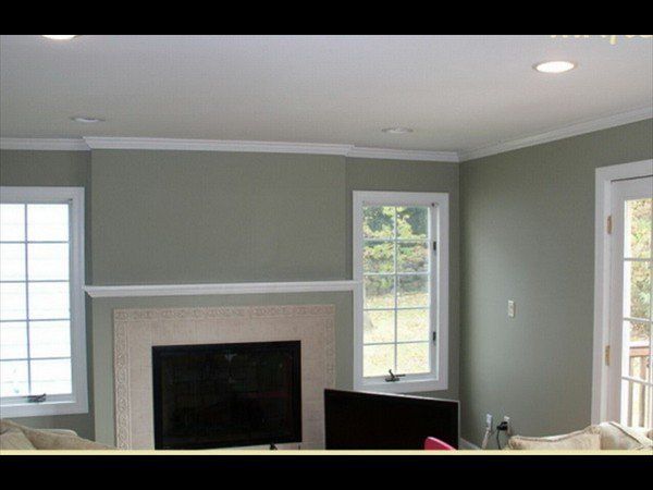 A living room with light gray-green walls, a fireplace with a light-colored surround, and two windows.