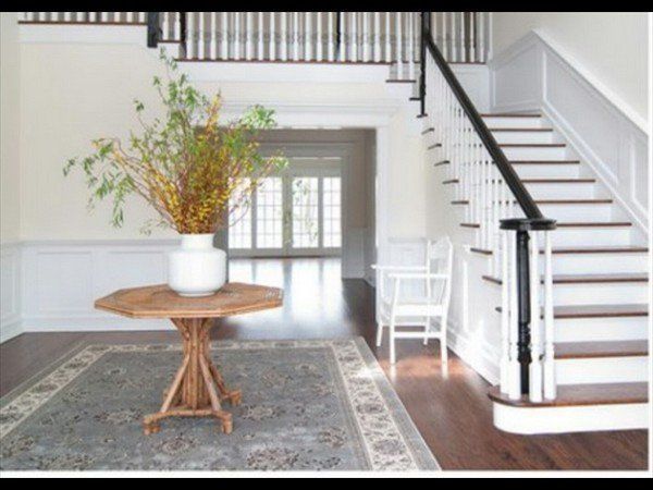A foyer features a central wooden octagonal table with a white vase of branches, set on a rug before a wooden staircase.