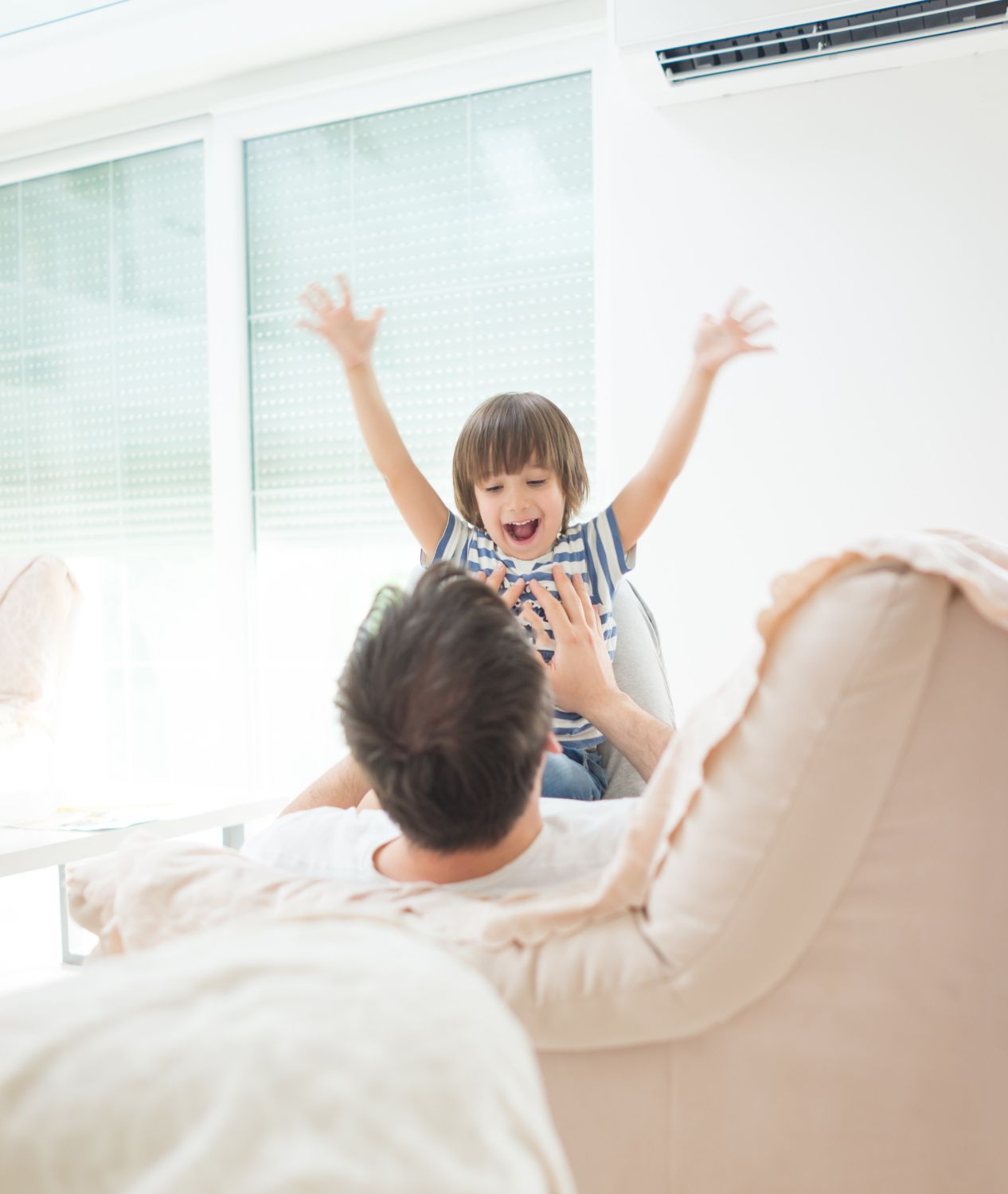 A child with arms raised happily sits on a man's lap, both indoors near a window.