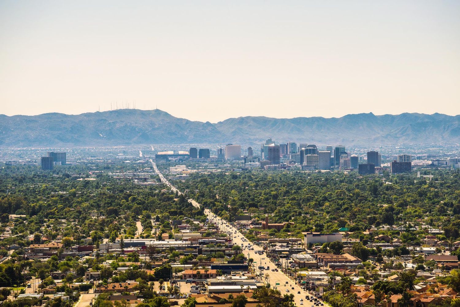 City skyline, with buildings rising above a green landscape, against a hazy mountain backdrop.