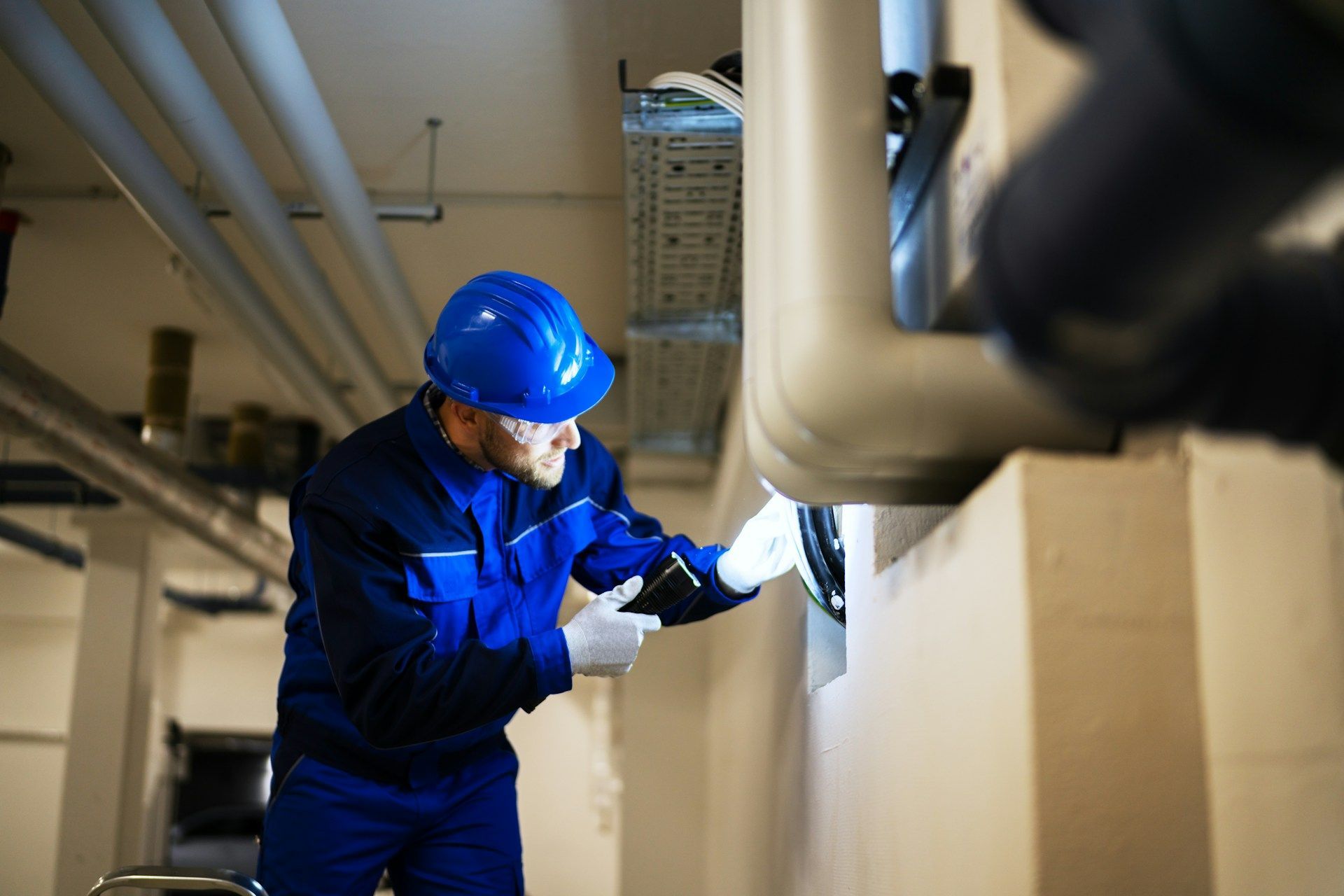 Technician in blue workwear inspects machinery with a flashlight in a mechanical room, wearing a hard hat.