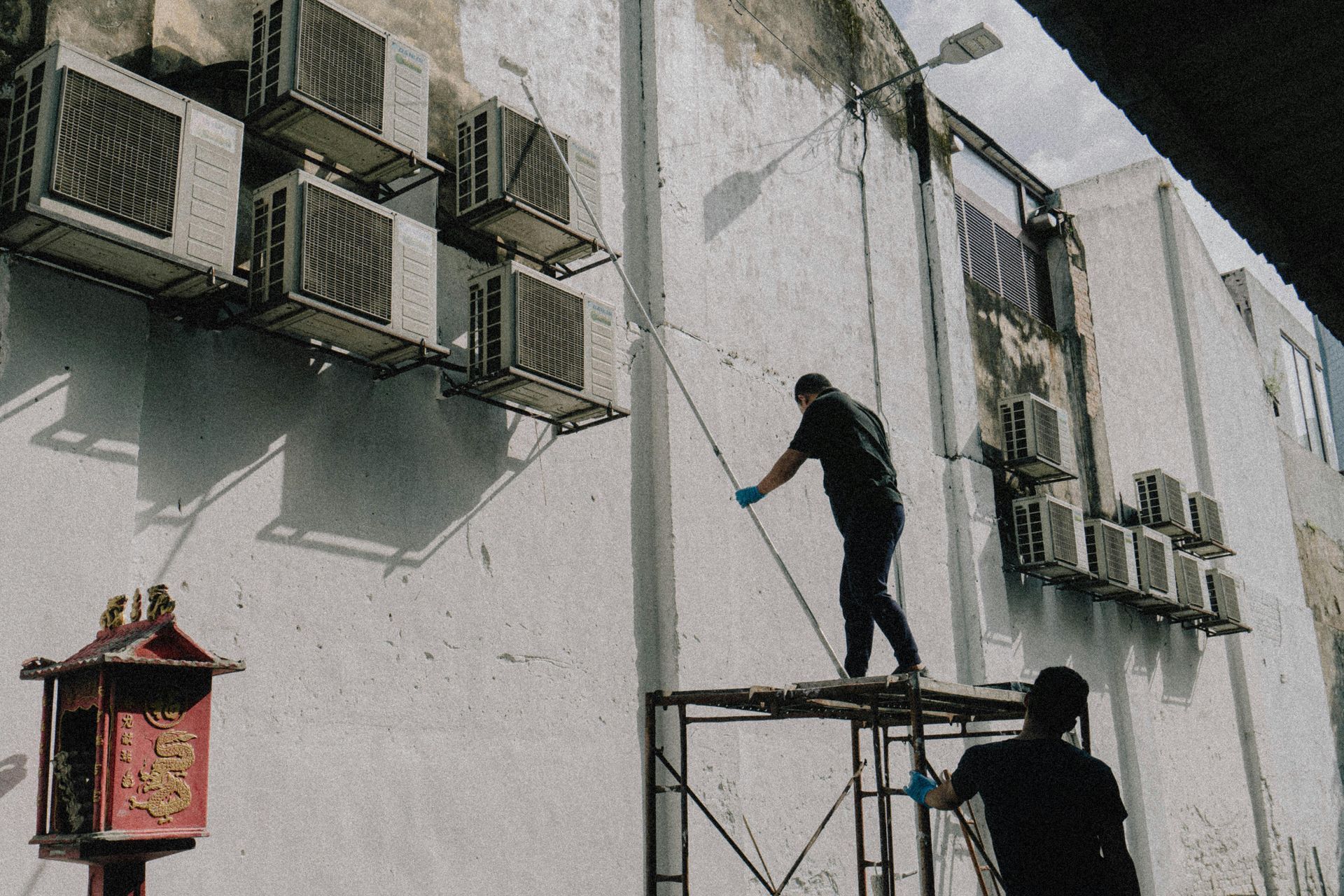 Two people cleaning a building exterior. One on scaffolding, spraying. Multiple AC units on wall.