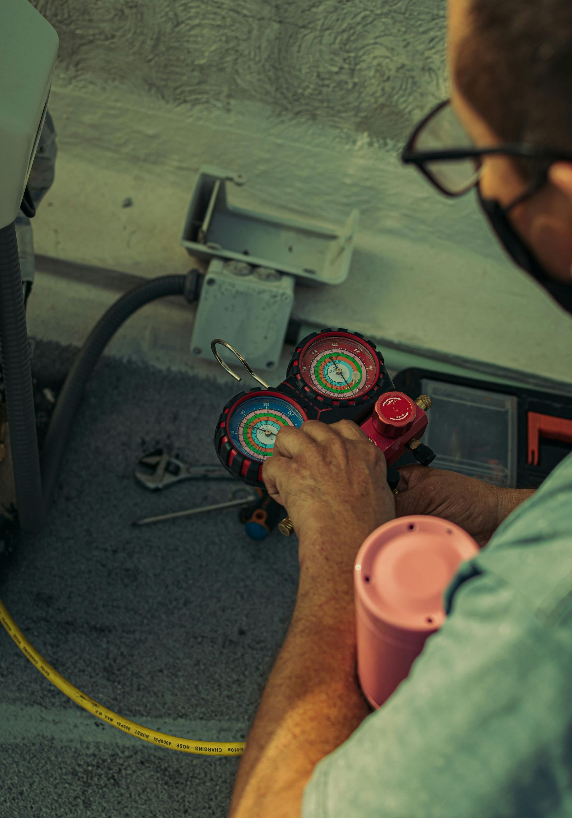 Person wearing glasses and a mask using gauges on a unit; yellow hose and pink container visible.
