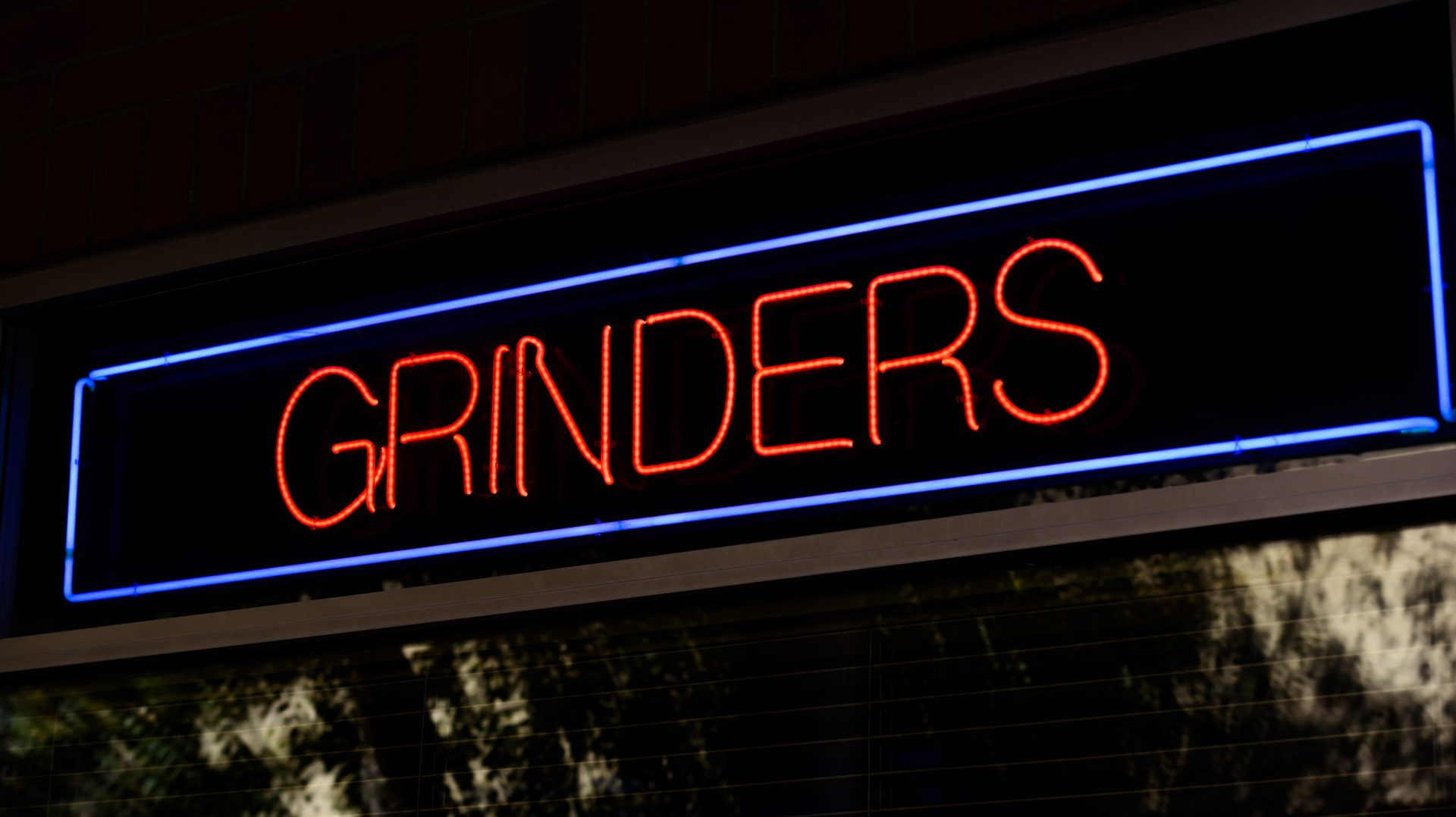 Neon “GRINDERS” sign in red letters outlined in blue against a dark background