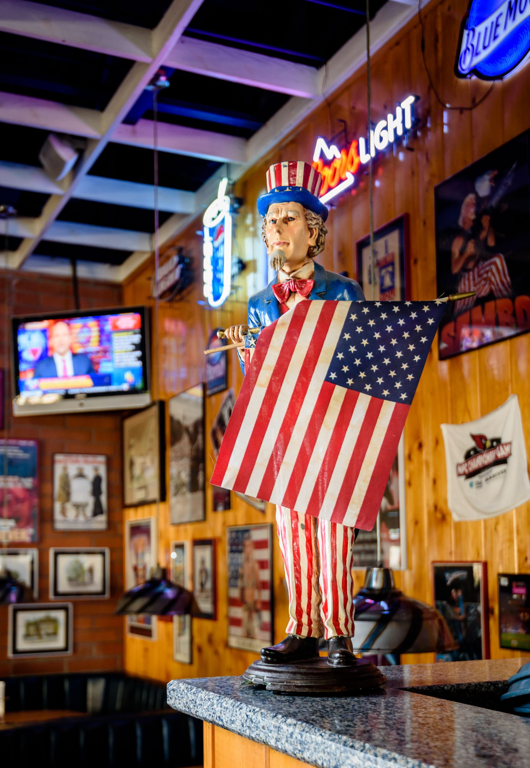 Life-sized Uncle Sam statue draped in an American flag inside a bar or restaurant with neon signs.
