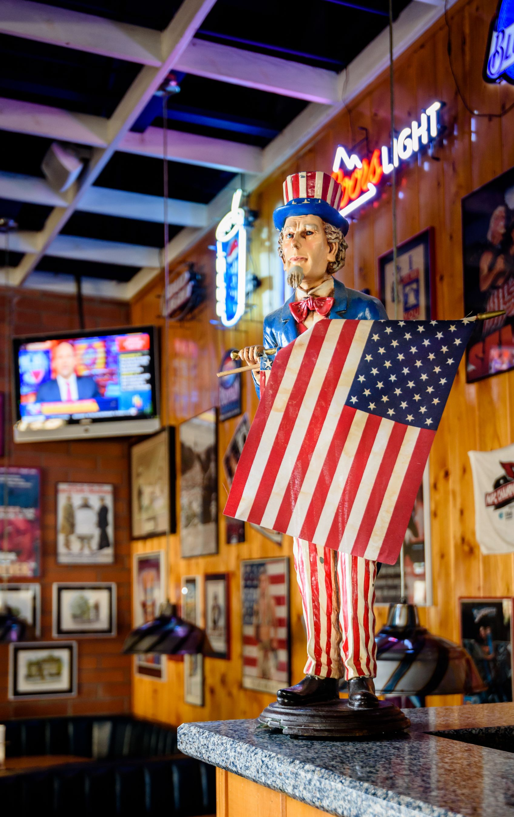 Bobblehead holding an American flag in a rustic bar with neon signs and framed wall art.