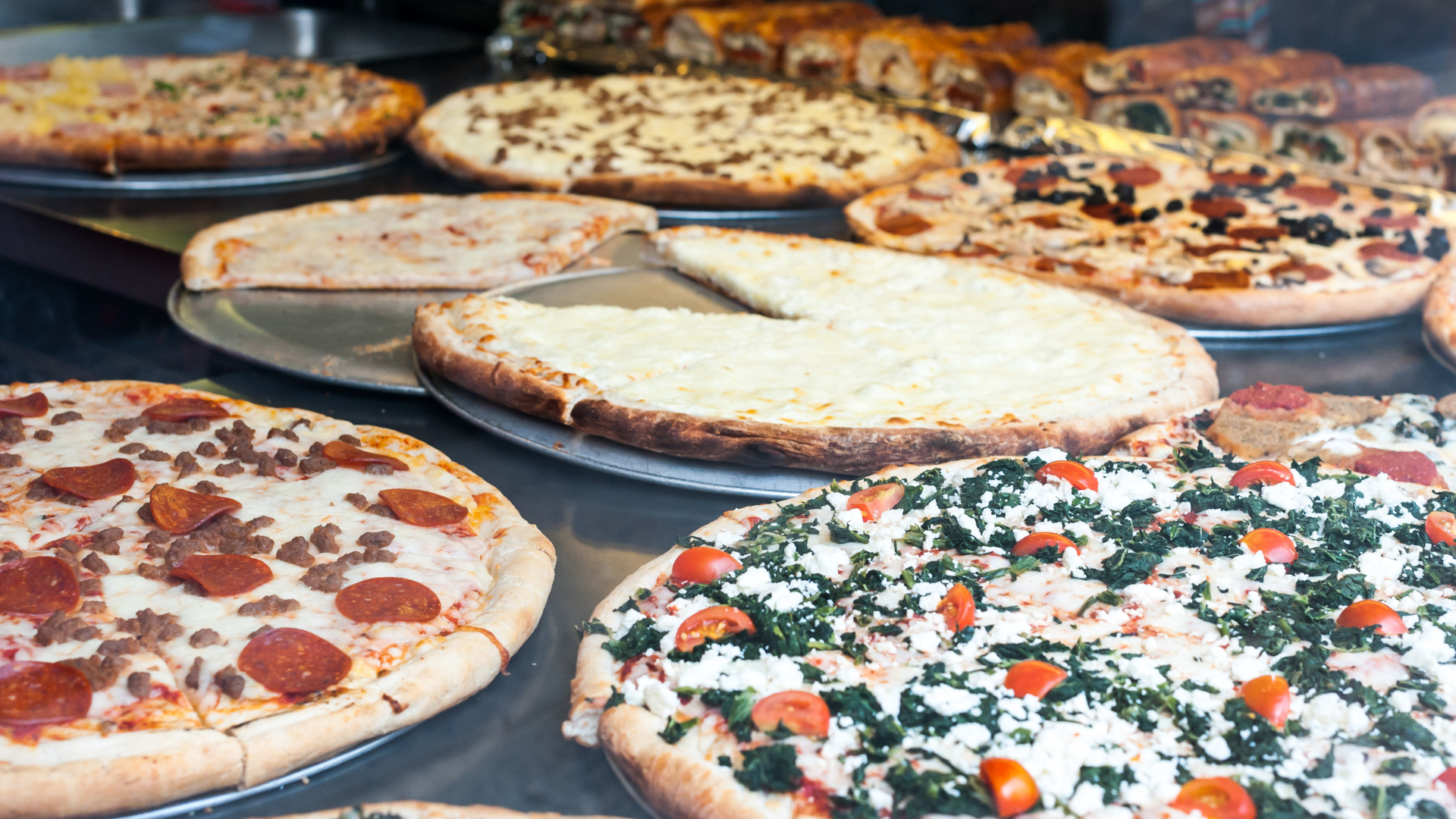 Assorted pizzas on trays, including pepperoni, cheese, and vegetable toppings, displayed at a buffet.