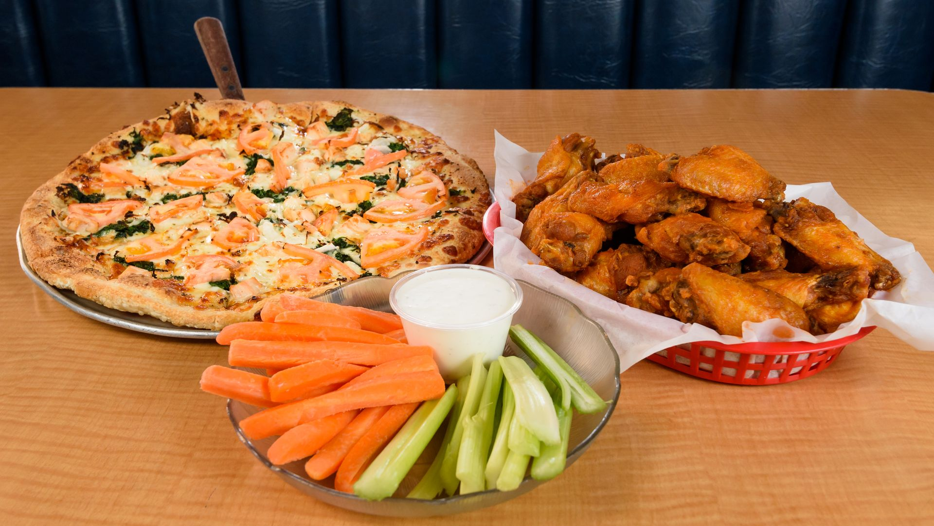Pizza, chicken wings, and vegetable sticks with dip on a table against a blue backdrop