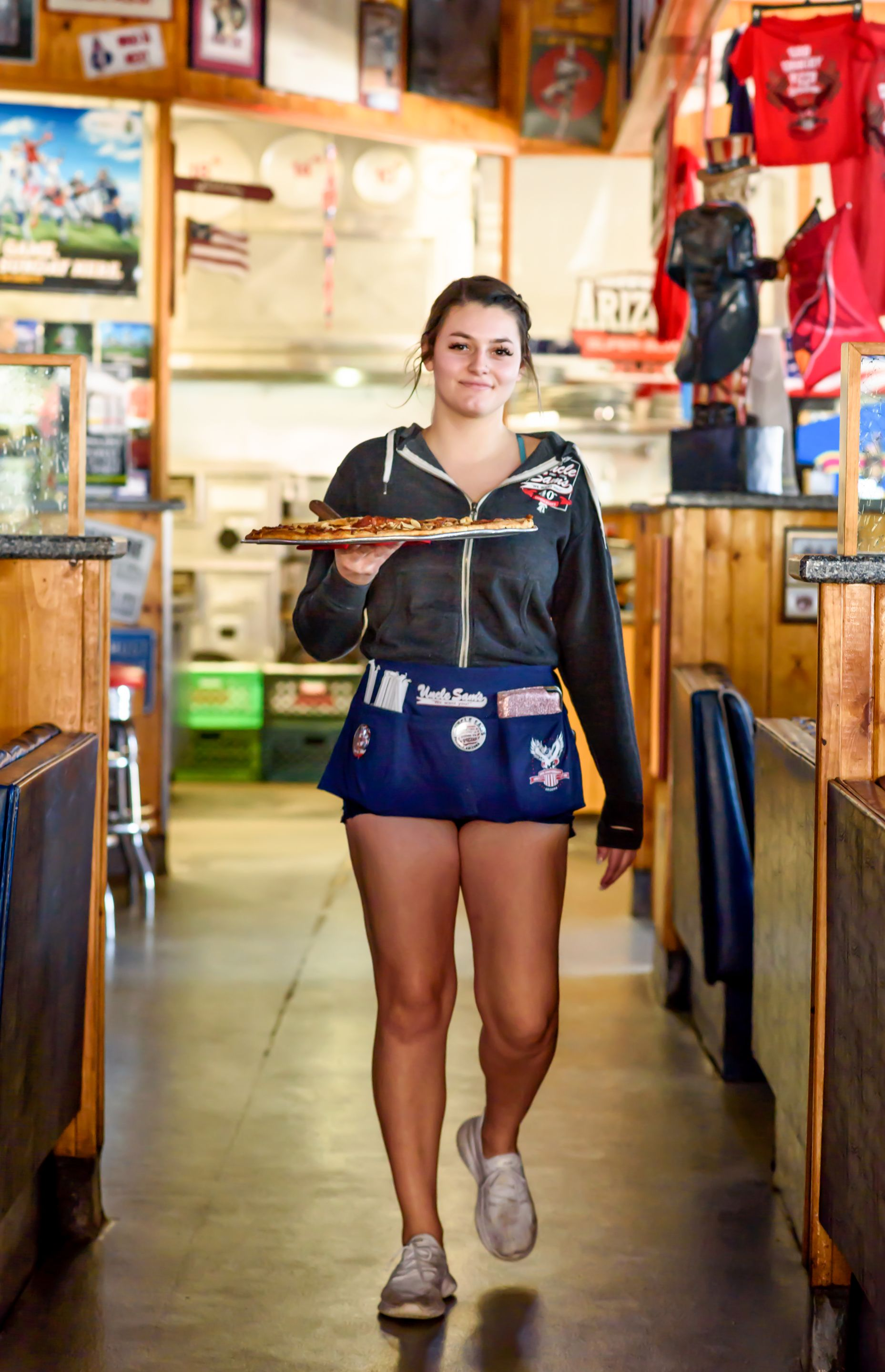 Waitress carrying a tray in a rustic café, wearing a white shirt and blue shorts, walking toward the camera.