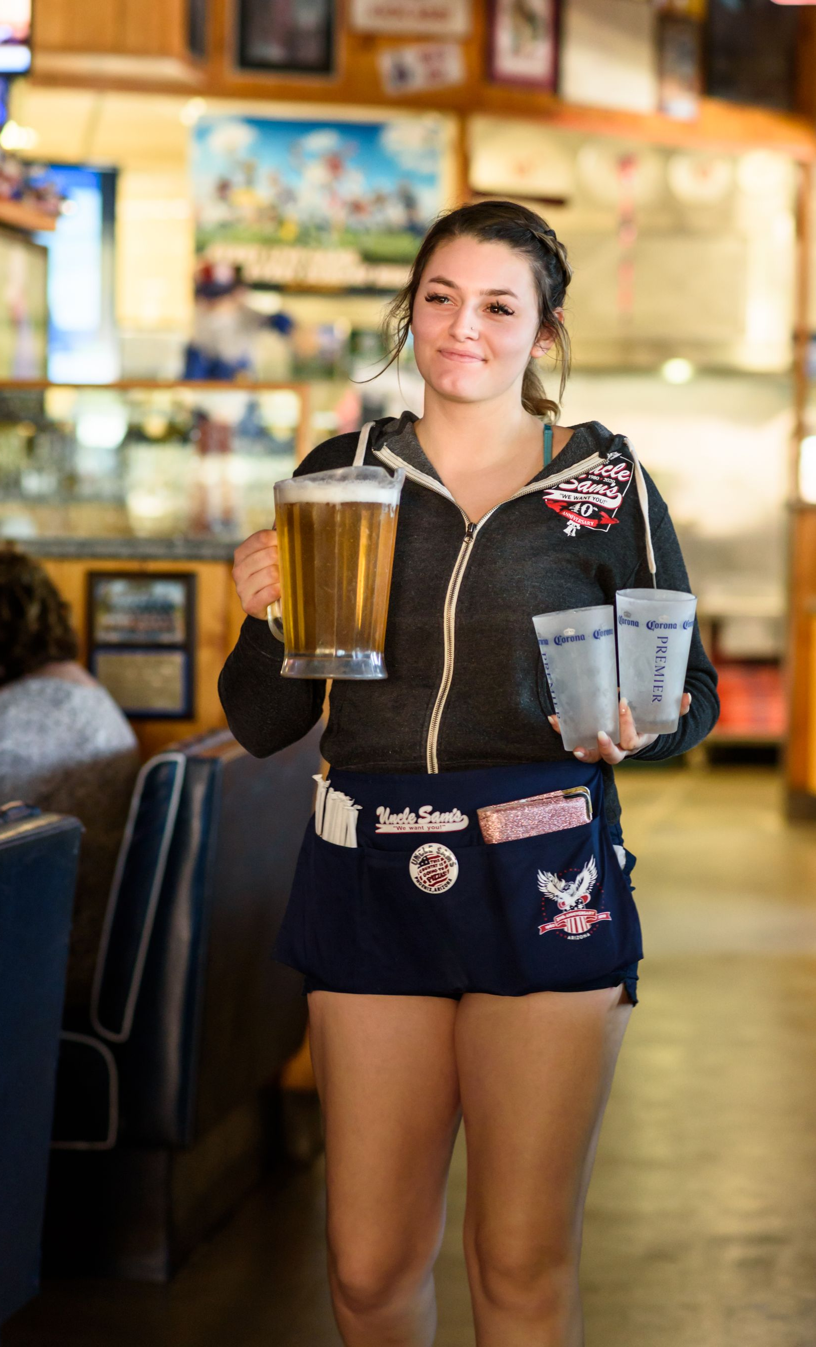 Waitress carrying two drinks in a restaurant, wearing a black top and dark shorts, smiling at the camera