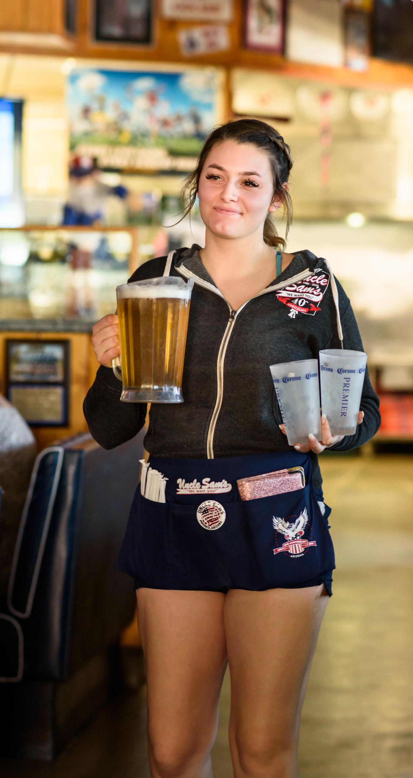 Waitress carrying two drinks in a restaurant, wearing a black top and dark shorts, smiling at the camera